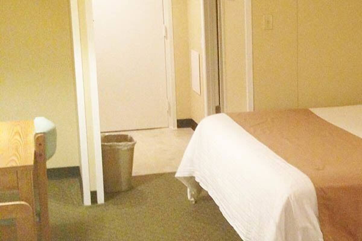 A hotel room with a bed, bedside table, chair, and a trash bin.