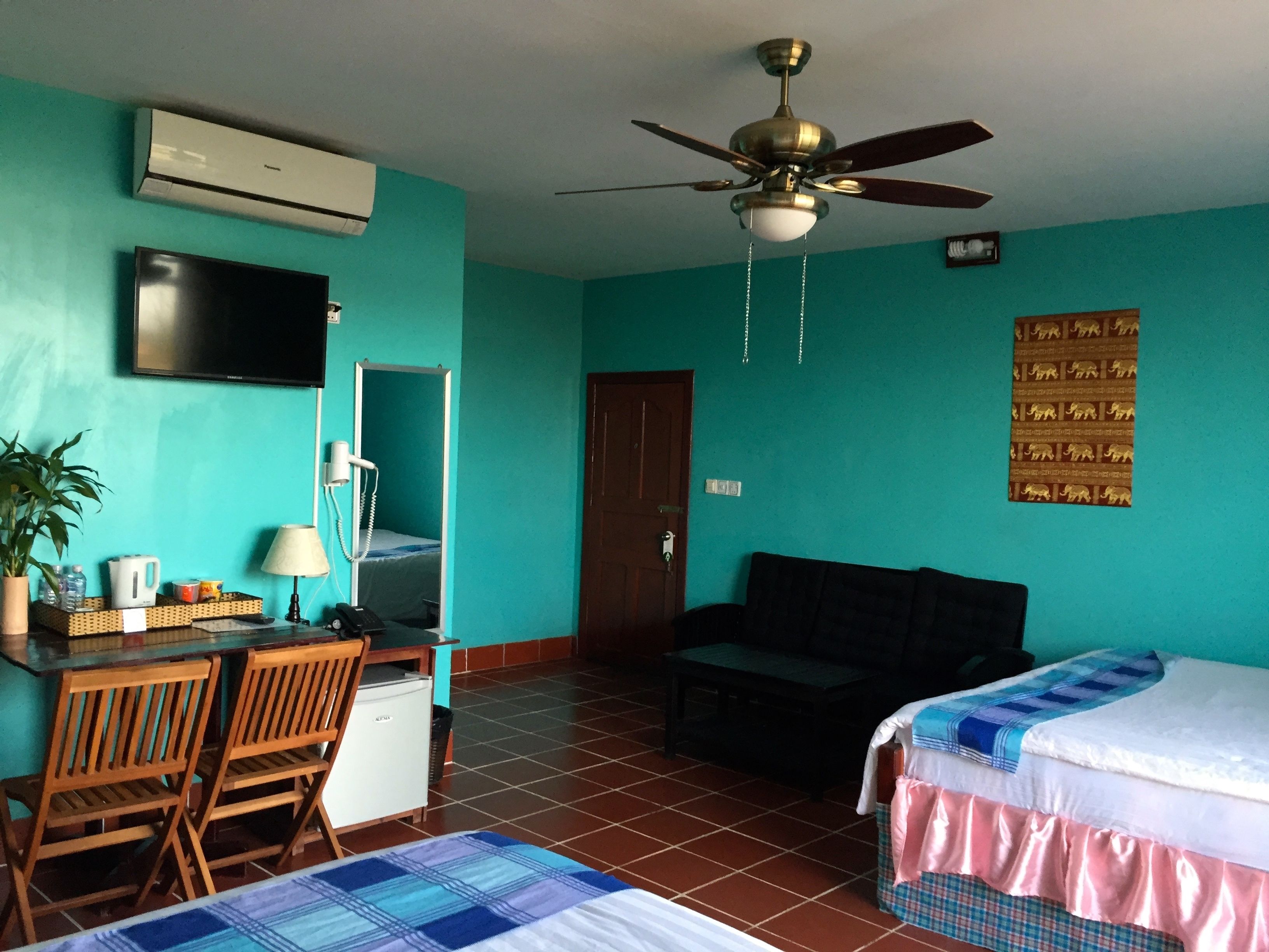 A room with teal walls, a ceiling fan, a TV, a desk with a lamp, a chair, a bed, and a refrigerator.