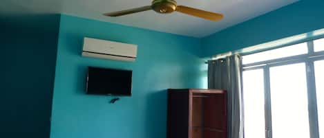 Una stanza con pareti color acquamarina, una ventola a soffitto, un condizionatore d'aria, una TV a schermo piatto e un armadietto in legno.