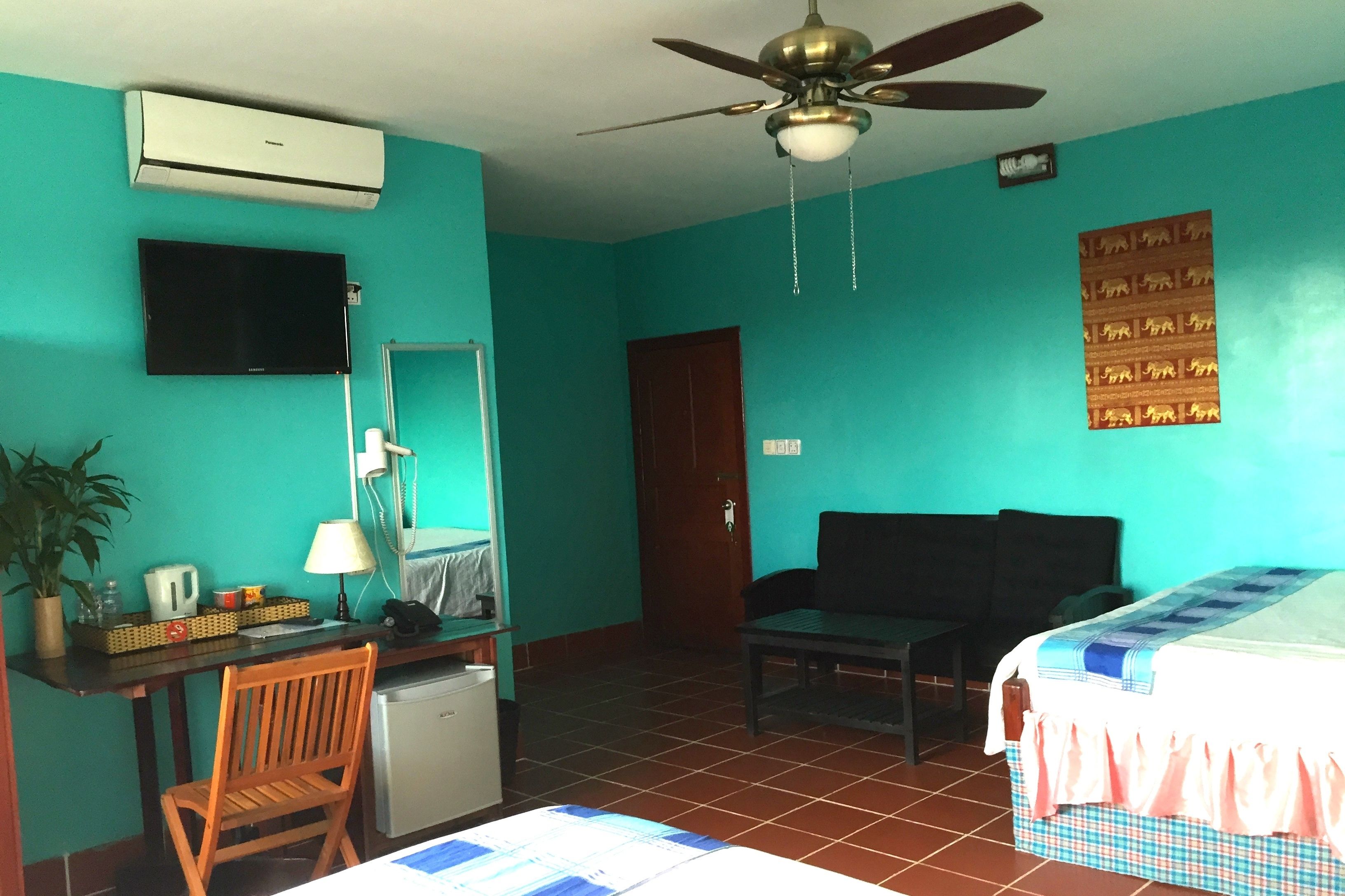 A room with teal walls, a ceiling fan, a bed with a checkered bedspread, a desk with a lamp, a chair, a small refrigerator, and a mounted TV.