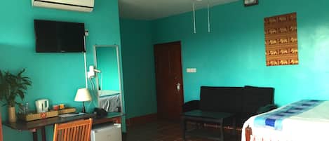 Ein Zimmer mit Türkiswänden, einem Deckenventilator, einem Bett mit kariertem Überwurf, einem Schreibtisch mit Lampe, einem Stuhl, einem kleinen Kühlschrank und einem Wand-Fernseher.