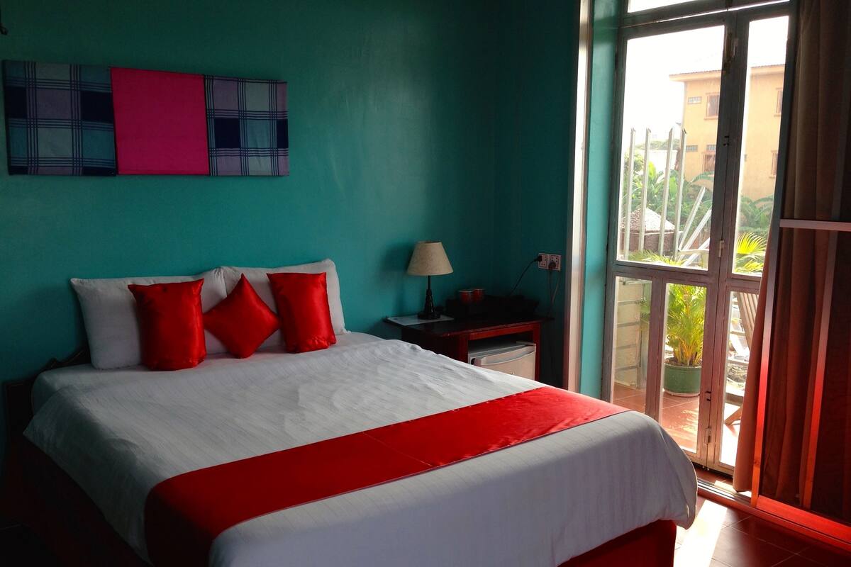 A hotel room with a bed, a nightstand, a lamp, and a view of the outside.