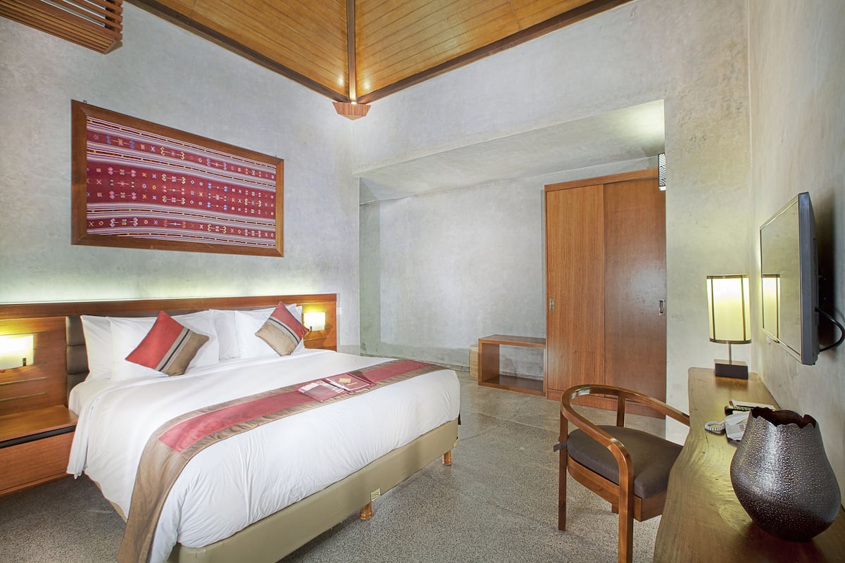 Habitación de hotel con una cama grande, cabecera de madera, mesitas de noche con lámparas, una silla de madera, una pequeña mesa con un jarrón y un tapiz de gran tamaño en la pared.
