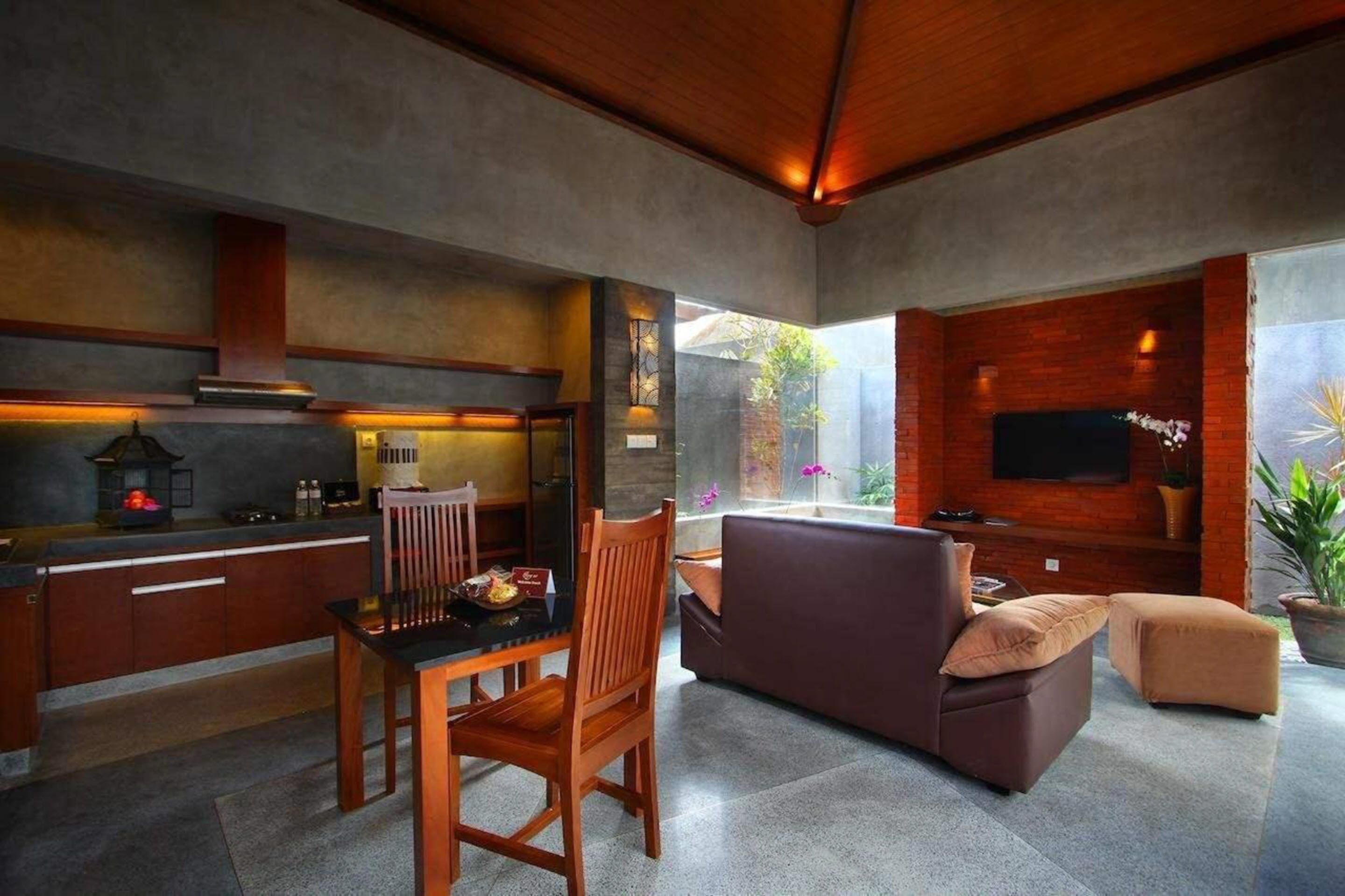 A modern kitchen with a dining area, a flat-screen TV mounted on a brick wall, and a comfortable seating area with a sofa and ottoman.