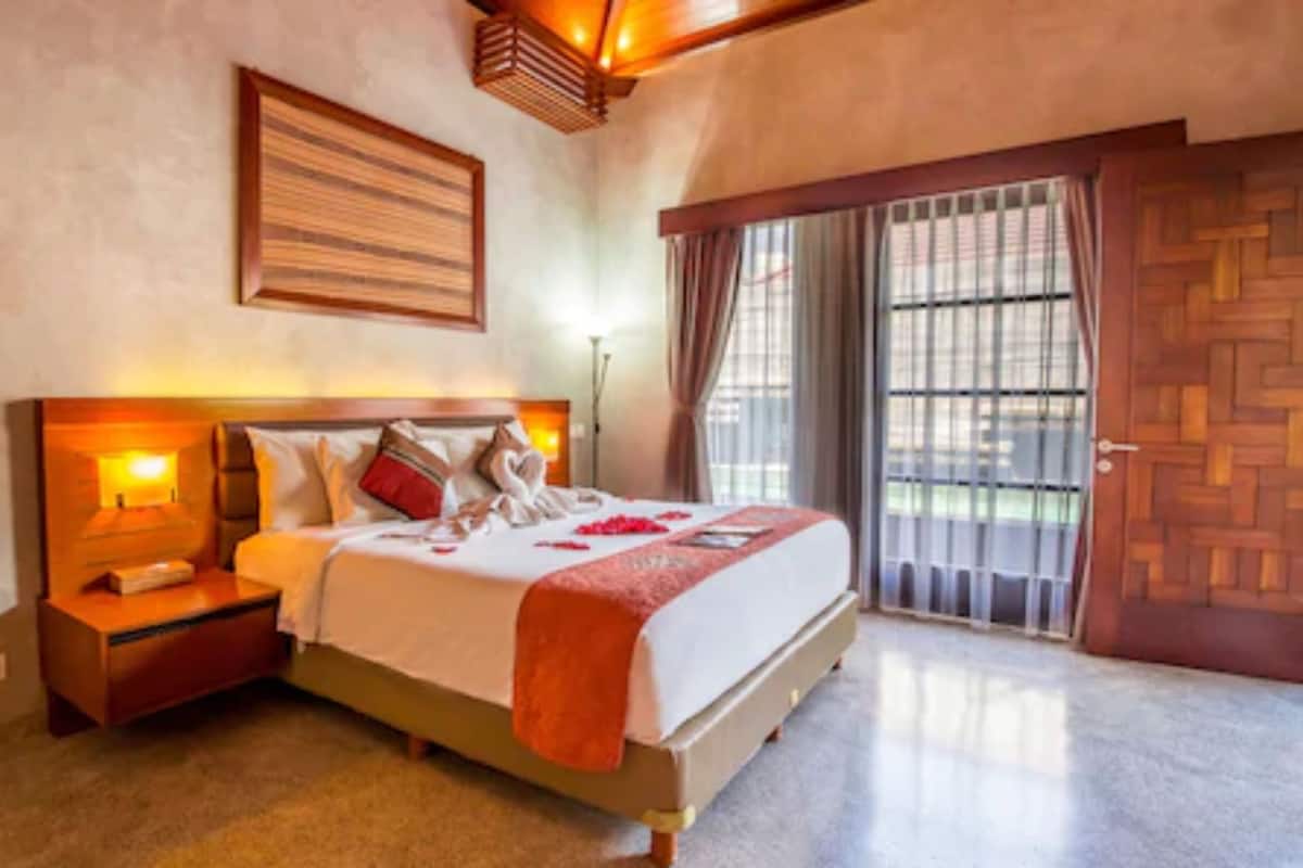 Una habitación de hotel con cama, mesita de noche, lámpara de pared, ventana con cortinas y puerta con diseño de madera.