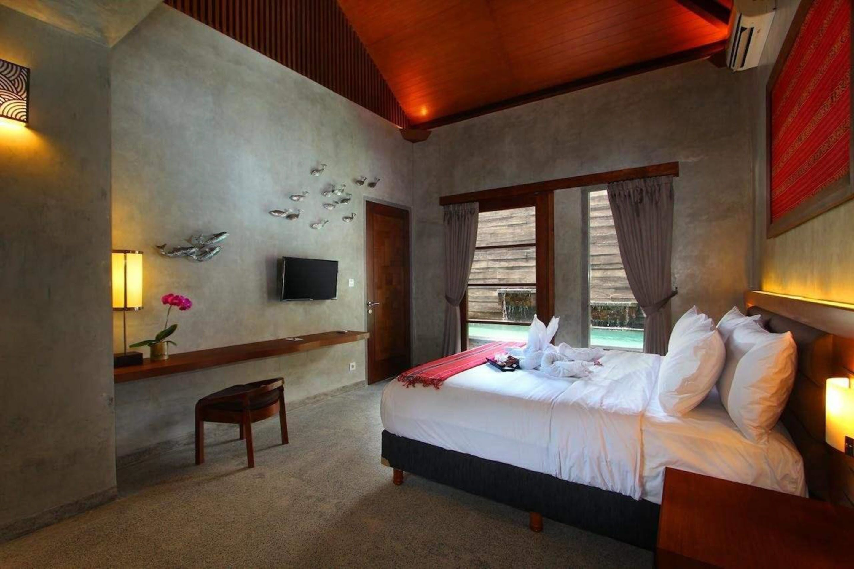 A bedroom with a bed, a TV, a small table with a lamp, a chair, and a window with curtains.