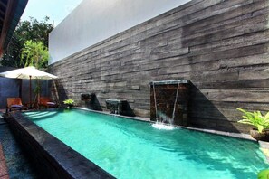 Una piscina con un muro de piedra de fondo, una fuente de agua y sillones de descanso.