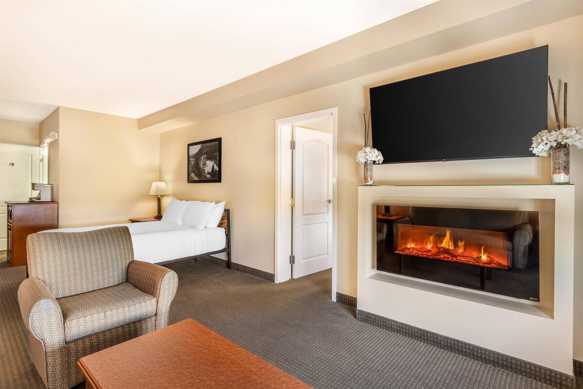 A hotel room with a bed, a sofa, a TV mounted above a fireplace, and a desk with a lamp.