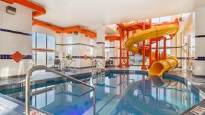 An indoor swimming pool with a yellow slide, clear poolside area, and a view of the outdoors through large windows.