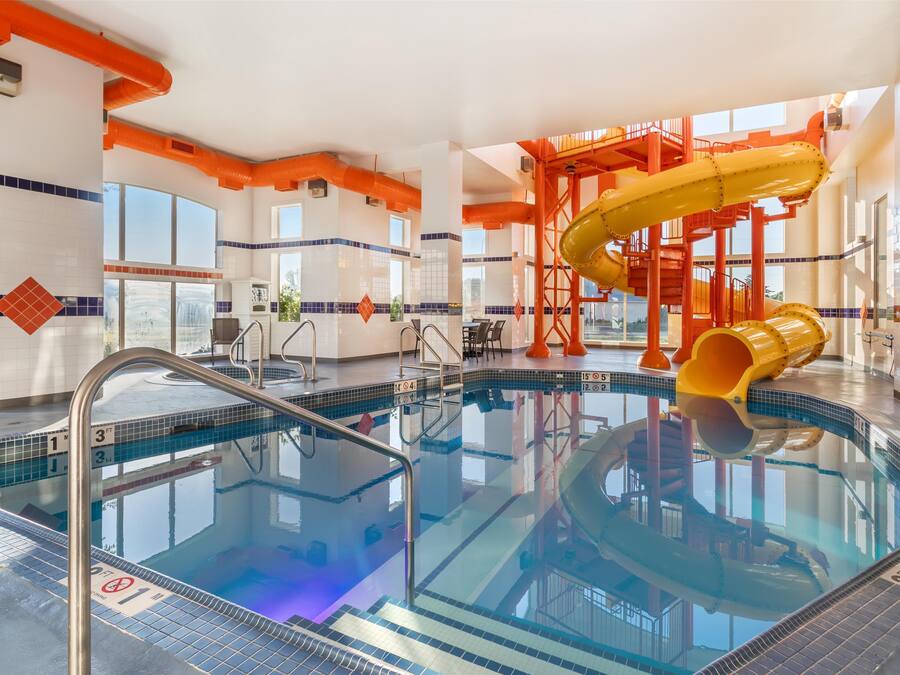 An indoor swimming pool with a yellow slide, clear poolside area, and a view of the outdoors through large windows.