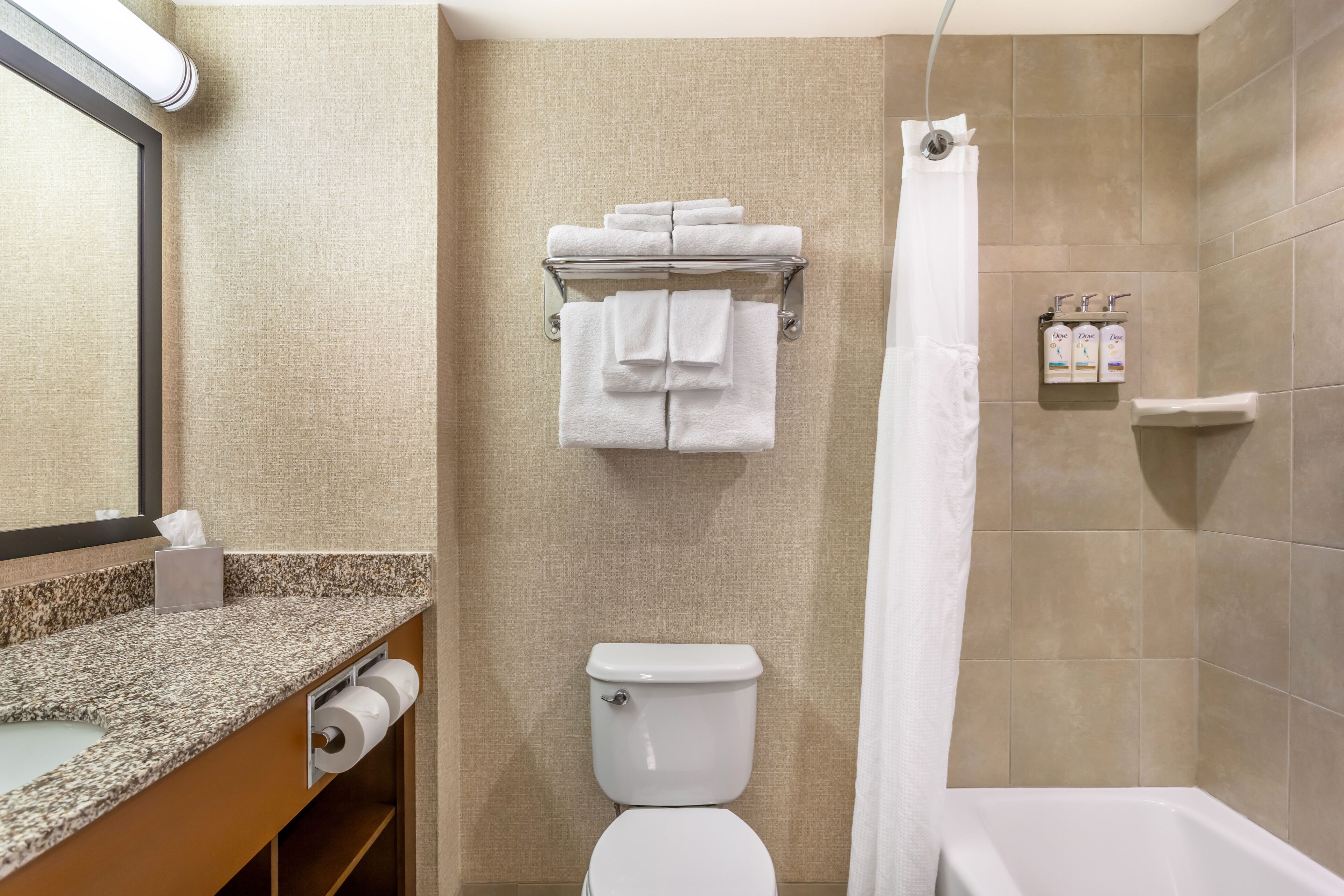 A bathroom with a toilet, a towel rack with folded towels, a shower curtain, a sink with a paper towel dispenser, and a soap dispenser on the wall.