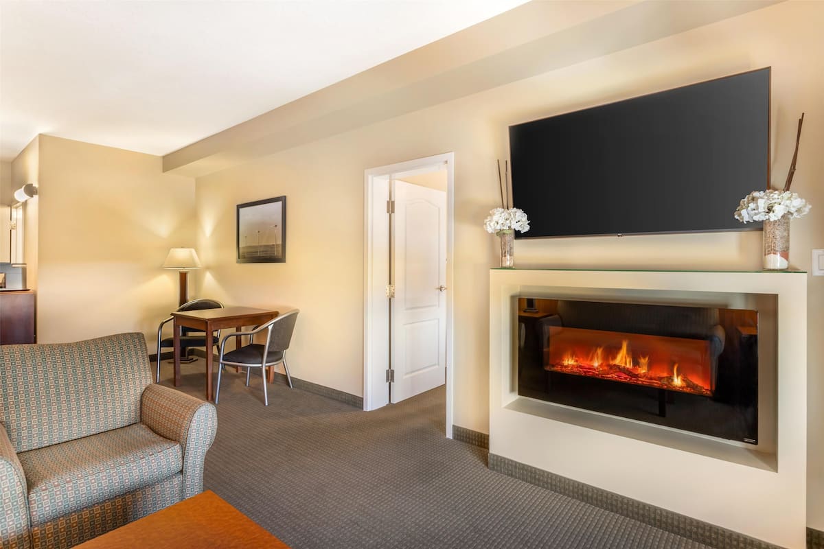 A hotel room with a sofa, a dining table with chairs, a flat-screen TV mounted above a fireplace, and a door leading to another room.