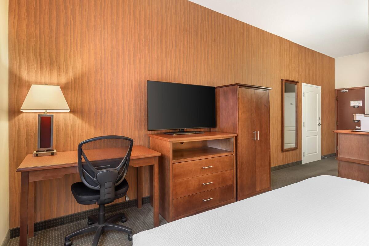 A hotel room with a wooden desk, a television, a chair, a bed, and a wardrobe.