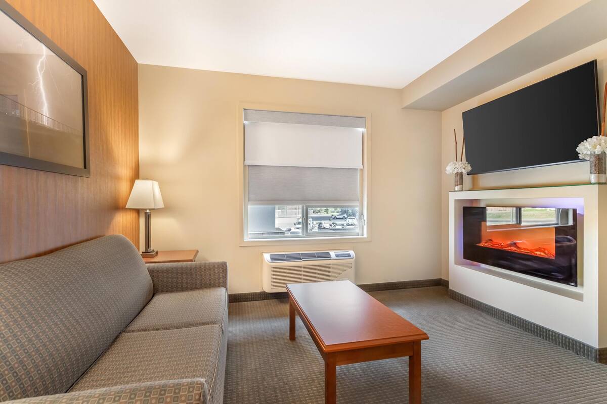 A hotel room with a sofa, a table, a TV, a fireplace, and a window with blinds.