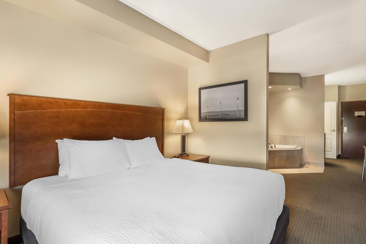 A hotel room with a large bed, a nightstand with a lamp, a painting on the wall, and a bathroom visible through an open door.