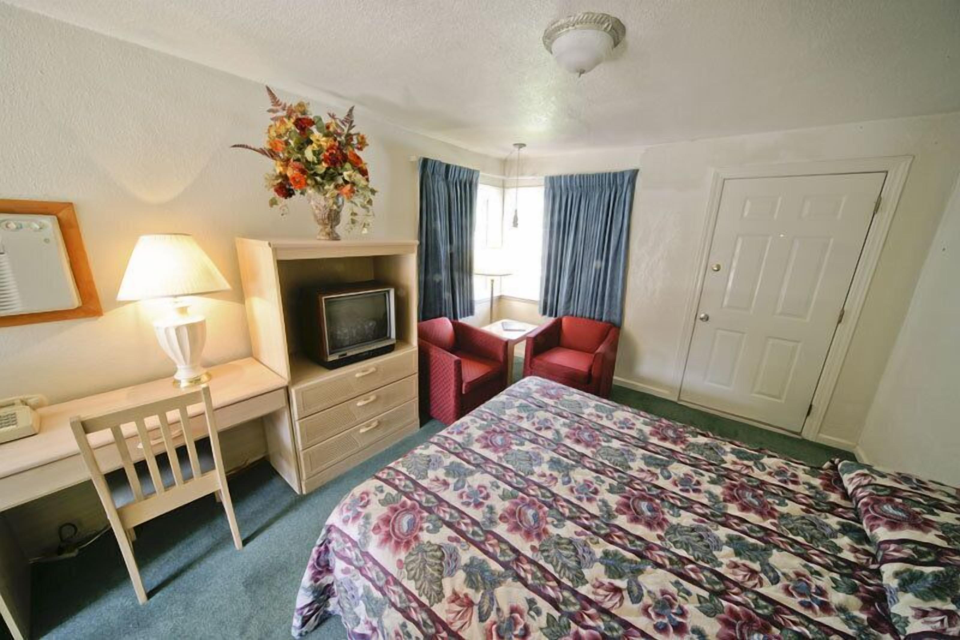 A hotel room with a bed, a desk with a lamp, a television, a chair, and a door.