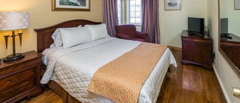 Un dormitorio ordenado con una cama grande, una mesita de noche de madera, un escritorio con televisor y un ventilador de techo.