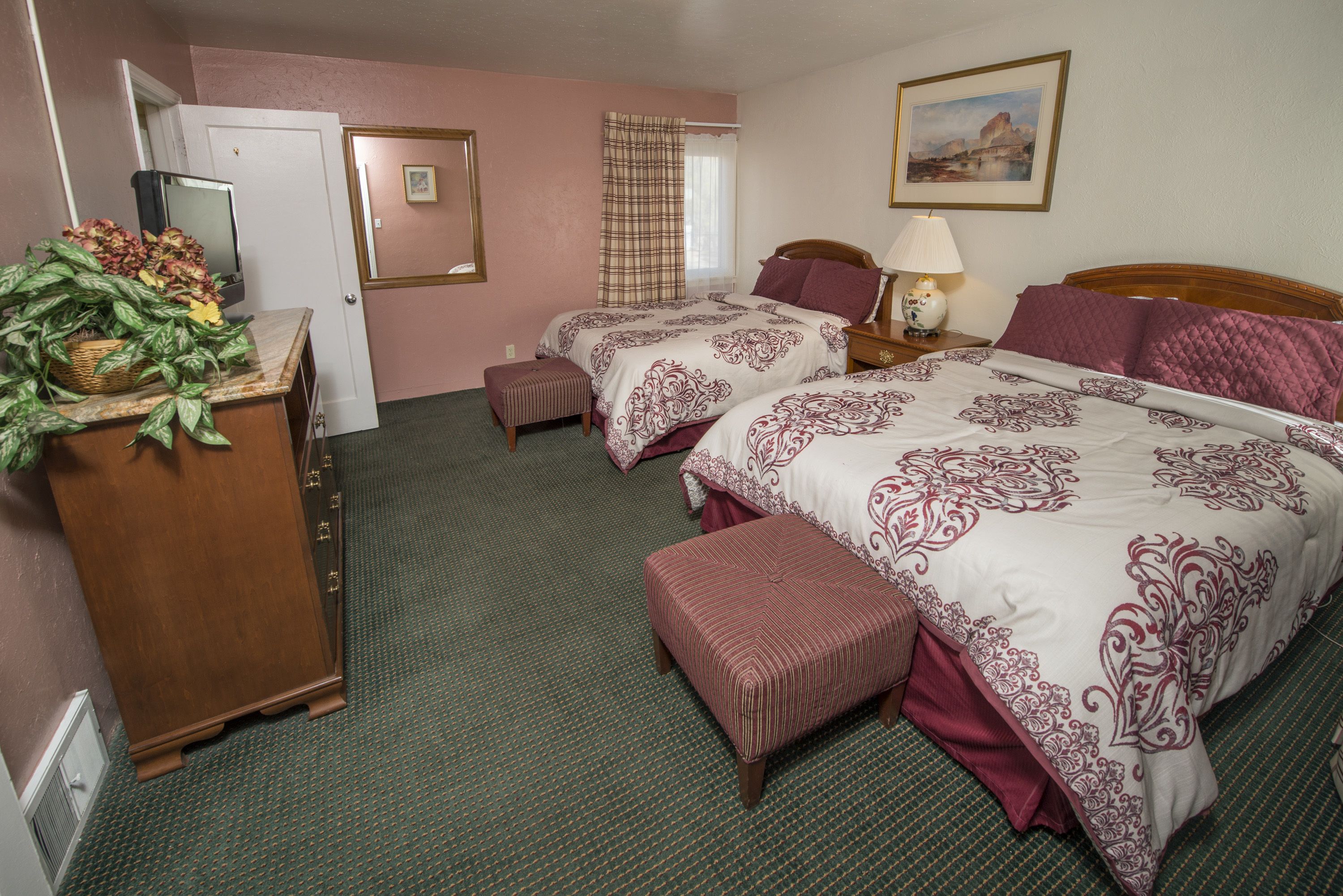 A hotel room with two beds, a wooden dresser, a desk with a computer, a window with curtains, and a painting on the wall.