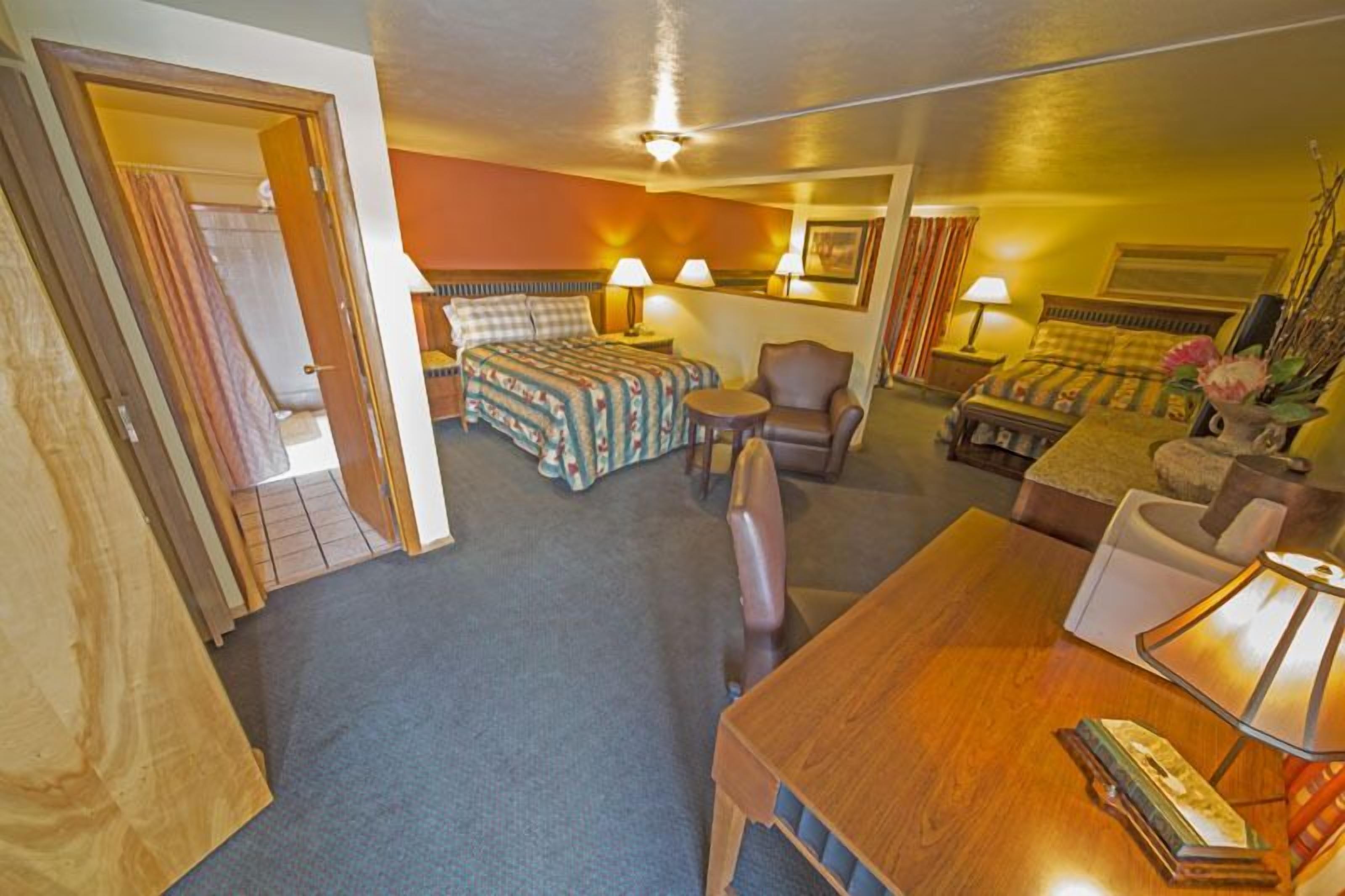 A hotel room with a bed, a desk, a chair, and a sofa. There are two lamps on the desk and a picture on the wall.