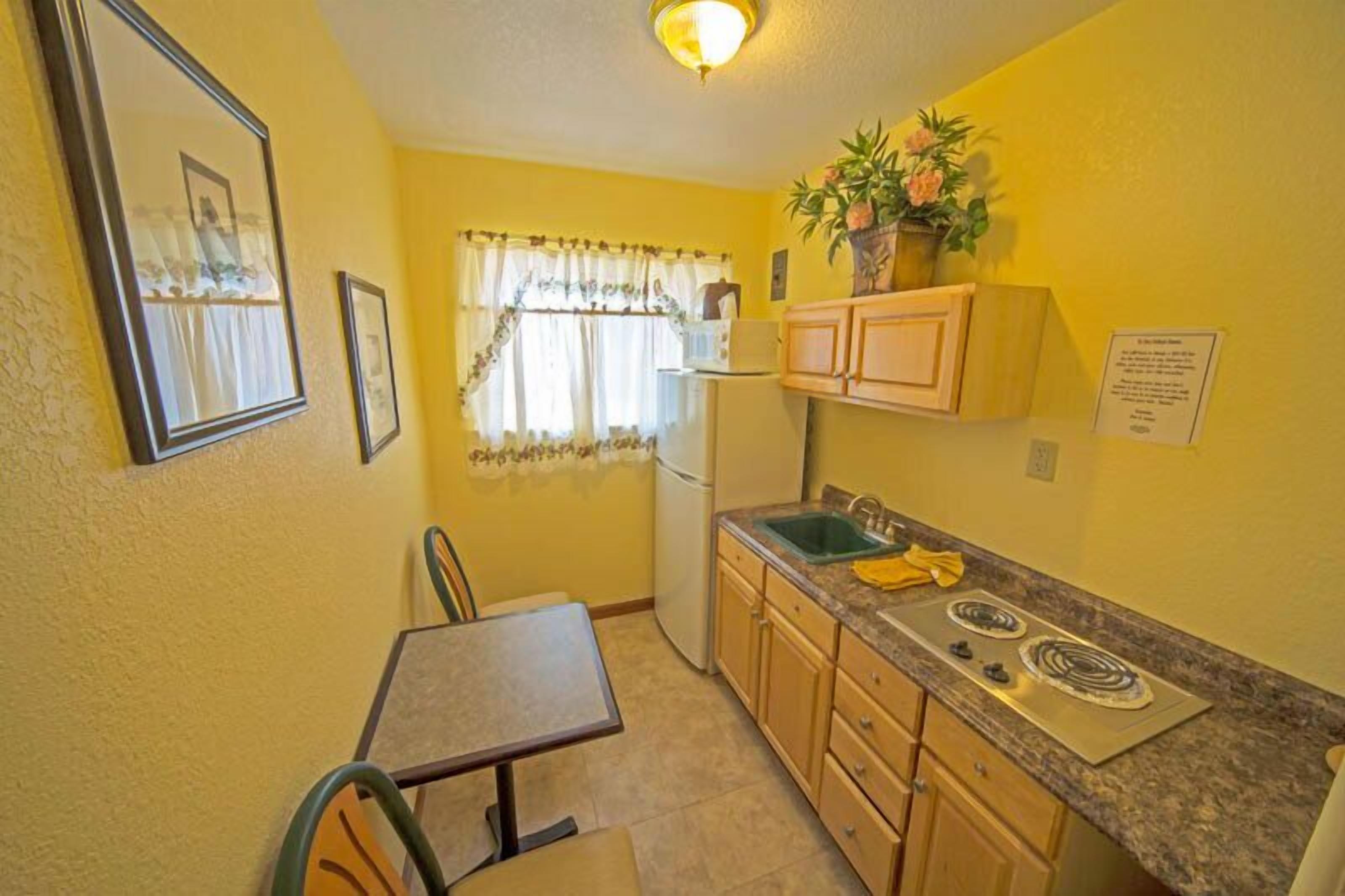 A compact kitchen with a refrigerator, stove, sink, and wooden cabinets. There is a dining table with chairs and a window with curtains.