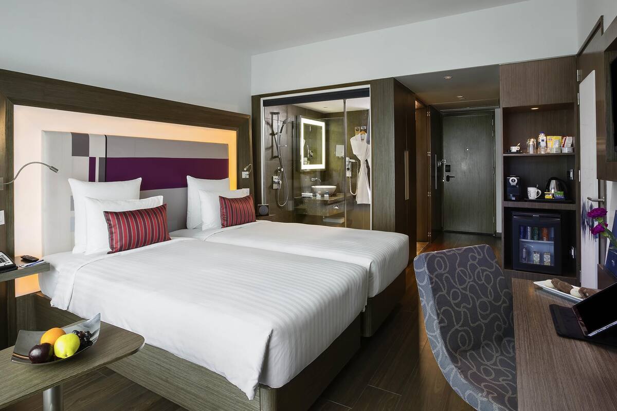 A hotel room with a large bed, a desk with a laptop, a glass shower area, and a minibar.