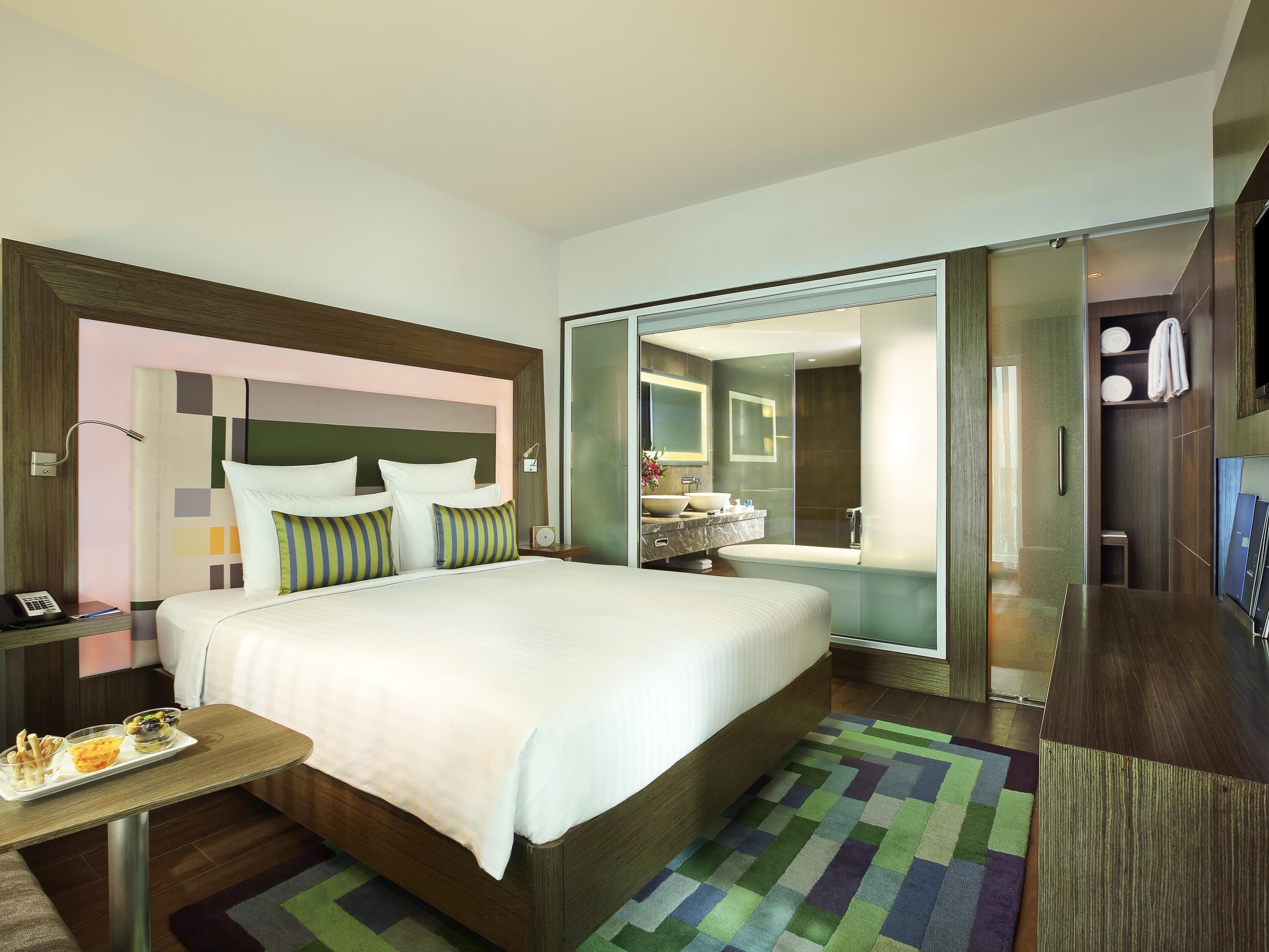 A hotel room with a bed, a bedside table with a phone and a tray of food, a bathroom with a sink and mirror, and a closet with hanging clothes.