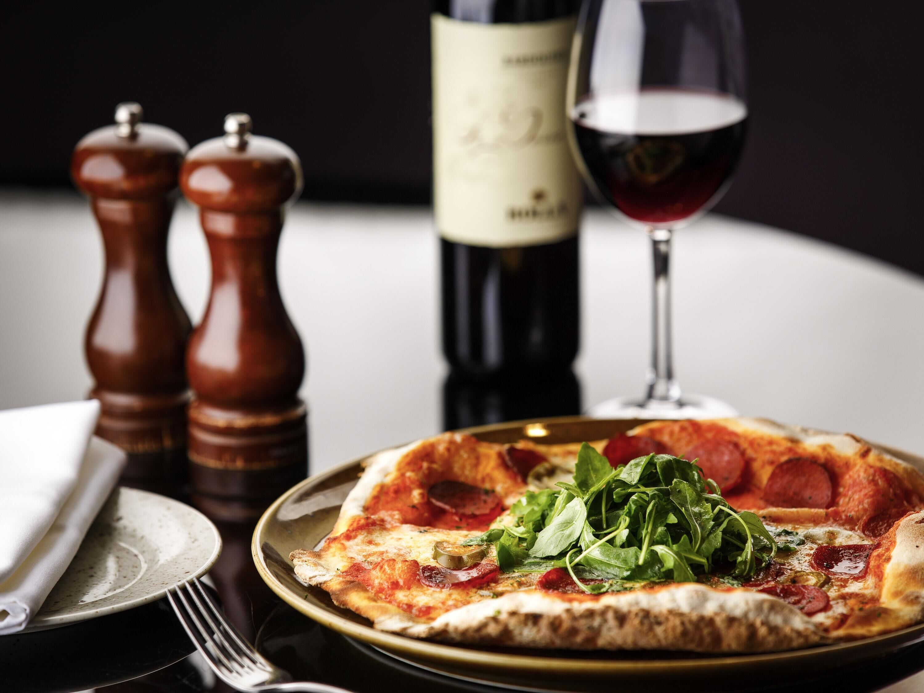 Una pizza con pepperoni y vegetales verdes en un plato, una botella de vino y dos saleros y pimenteros.