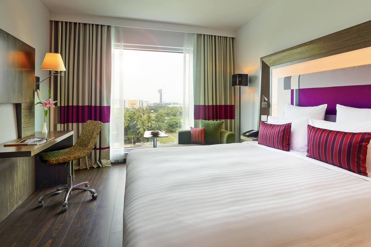 A hotel room with a large bed, a desk with a lamp, a chair, and a view of the cityscape.