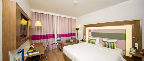 A hotel room with a large bed, a desk with a chair, a desk lamp, and a nightstand with a lamp.