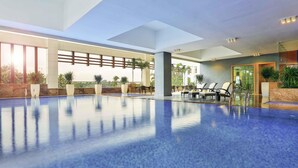 A large indoor swimming pool with clear blue water, surrounded by lounge chairs and potted plants. - Novotel Chennai Sipcot (Tirupporur)