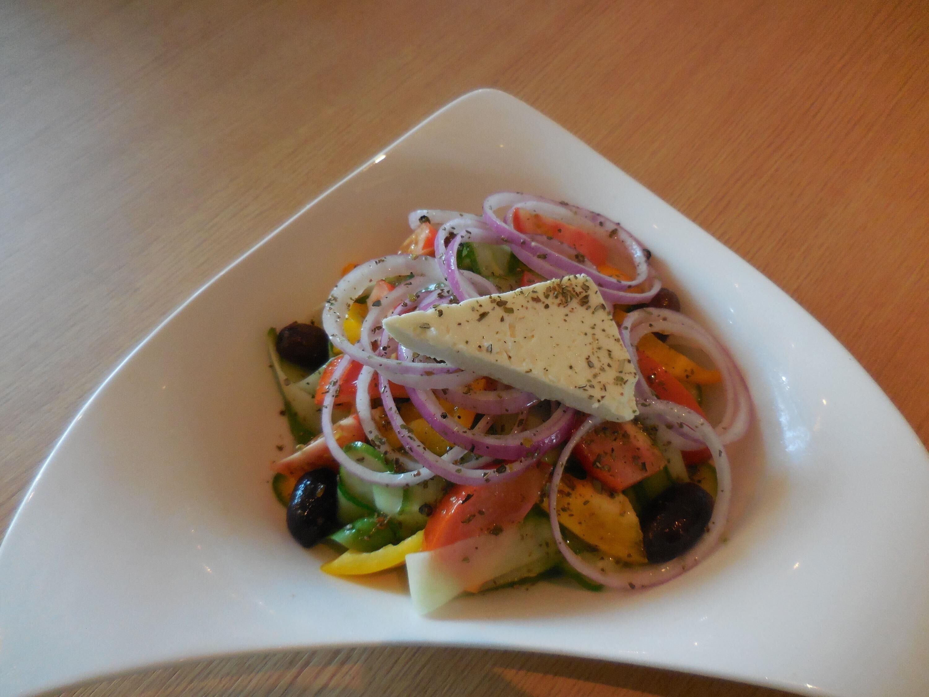 A white square plate with a salad containing tomatoes, cucumbers, red onions, black olives, and a piece of cheese on a wooden table.