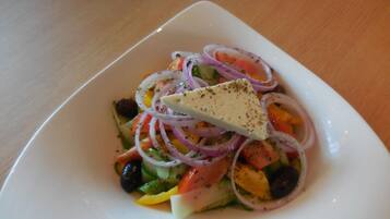 A white square plate with a salad containing tomatoes, cucumbers, red onions, black olives, and a piece of cheese on a wooden table.