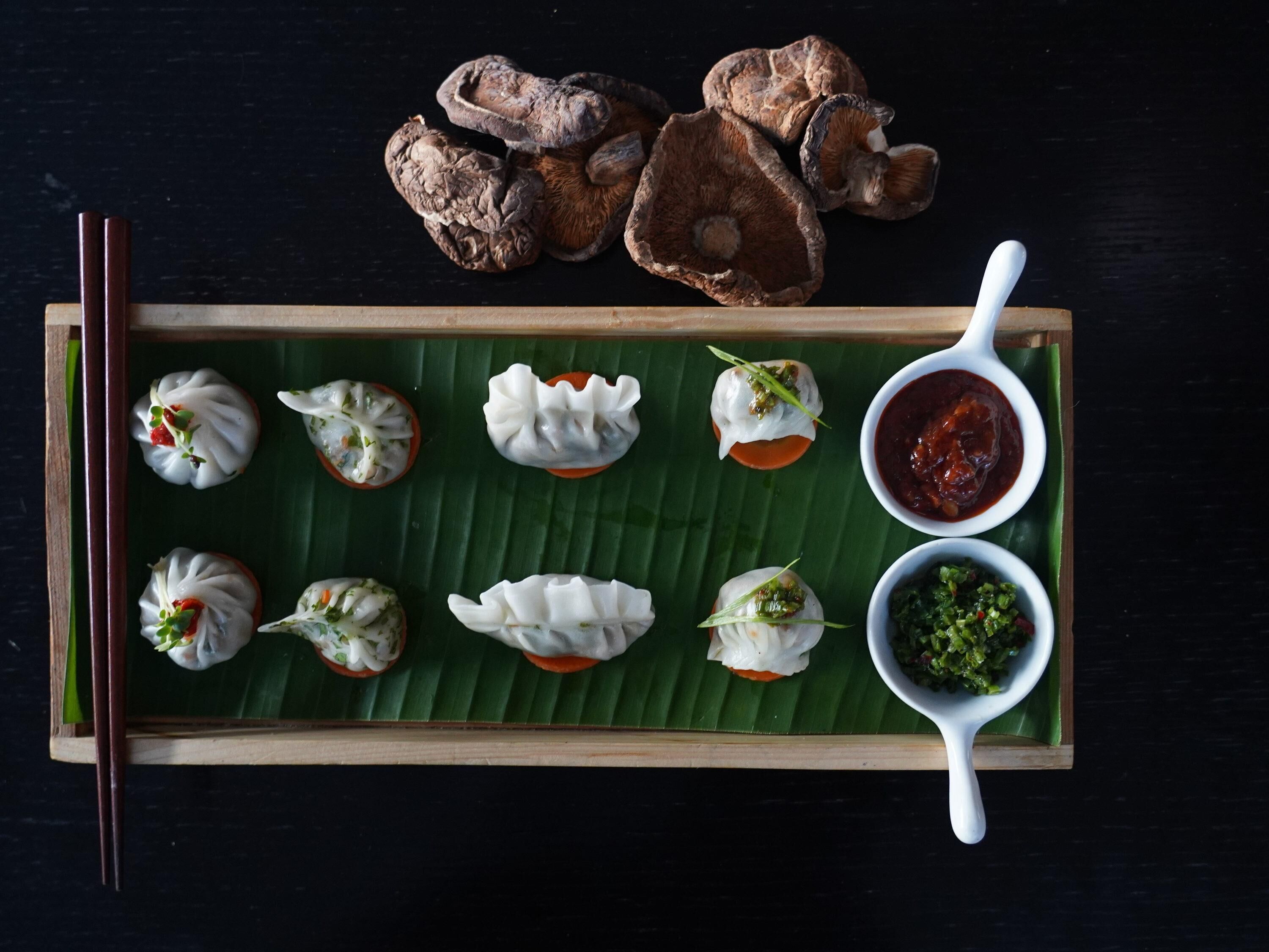 A wooden tray with dumplings on a banana leaf, accompanied by a spoon and two small bowls of dipping sauces.