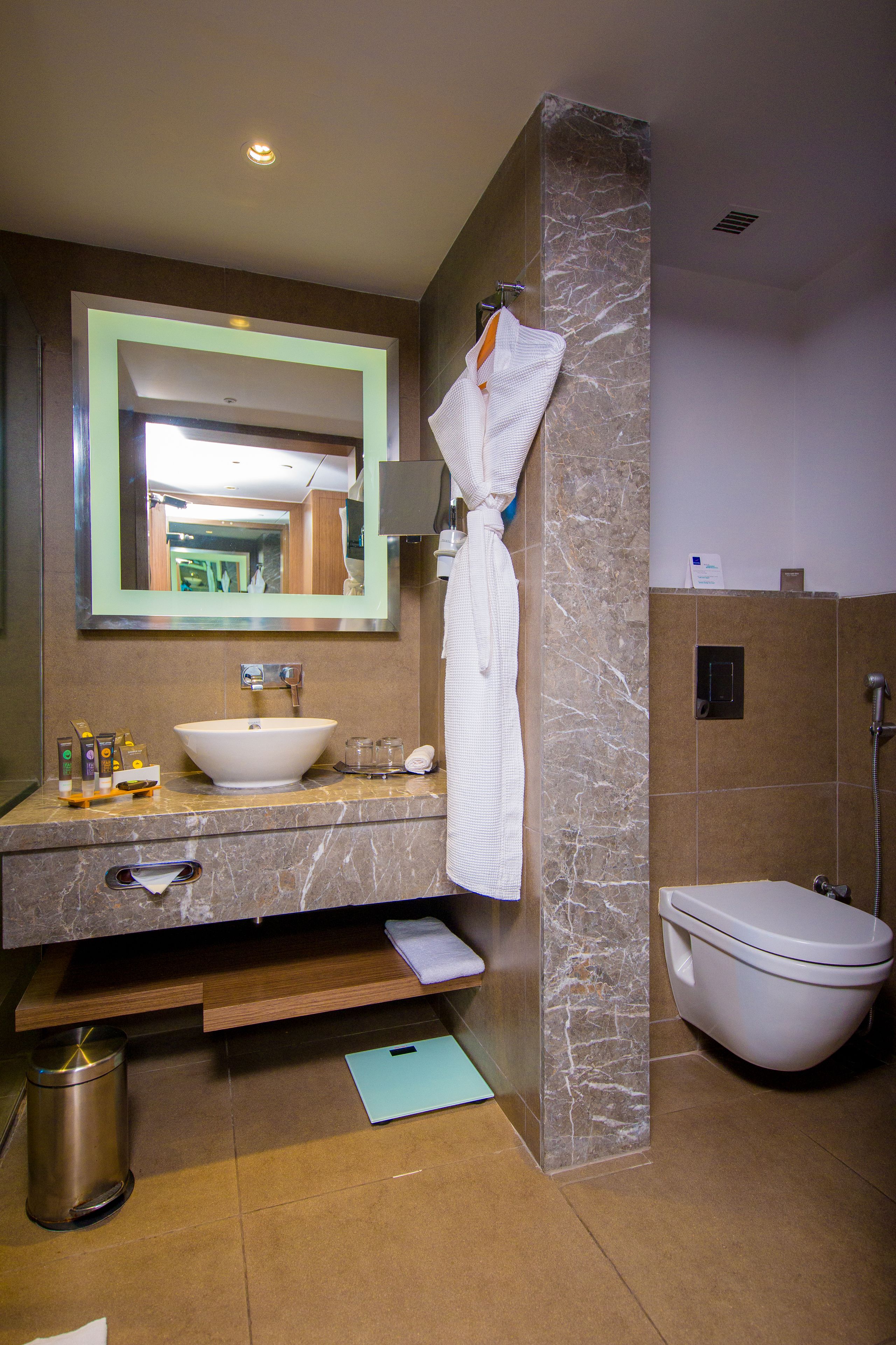 Un baño moderno con lavamanos de mármol, un espejo grande, una bata colgada en la pared y un área de inodoro separada.