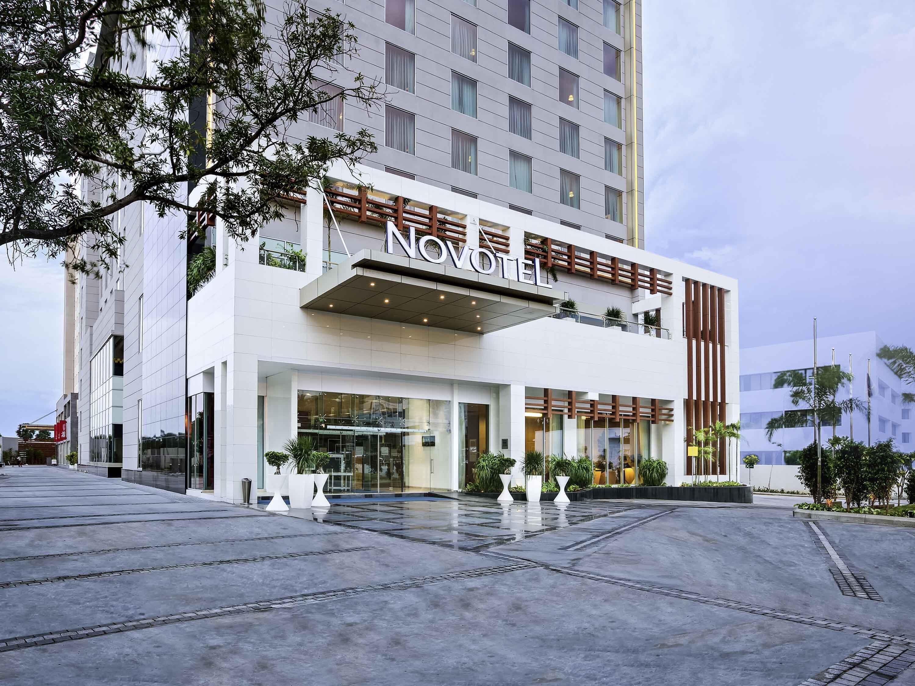 Un hotel moderno con un llamativo letrero 'NOVOMILE', un amplio acceso de vidrio y un espacioso área exterior con plantas en macetas.
