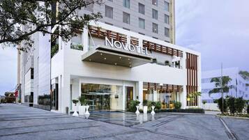Un hotel moderno con un llamativo letrero 'NOVOMILE', un amplio acceso de vidrio y un espacioso área exterior con plantas en macetas.