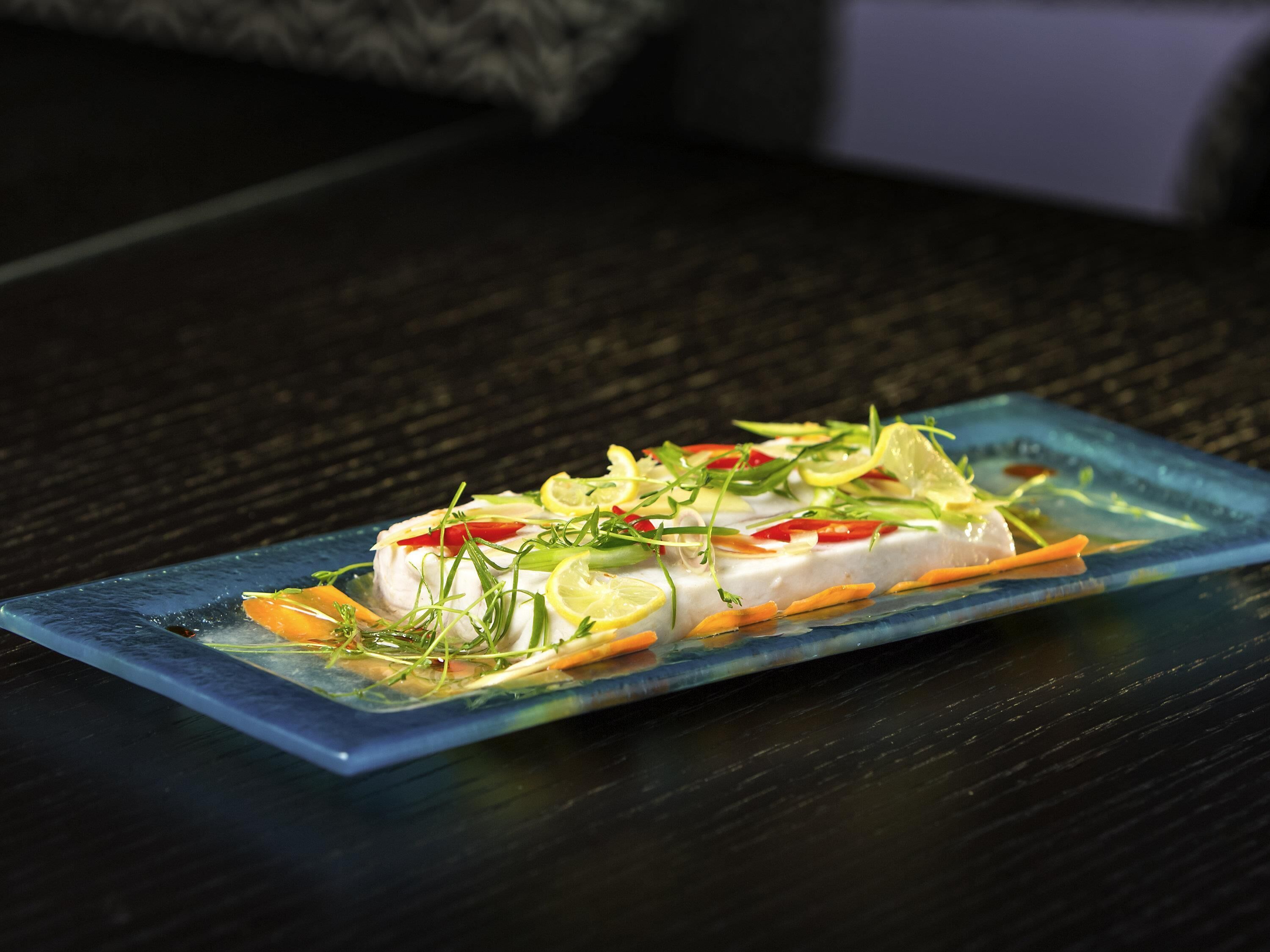 A rectangular glass plate with a white fish dish garnished with vegetables and herbs, placed on a dark textured surface.