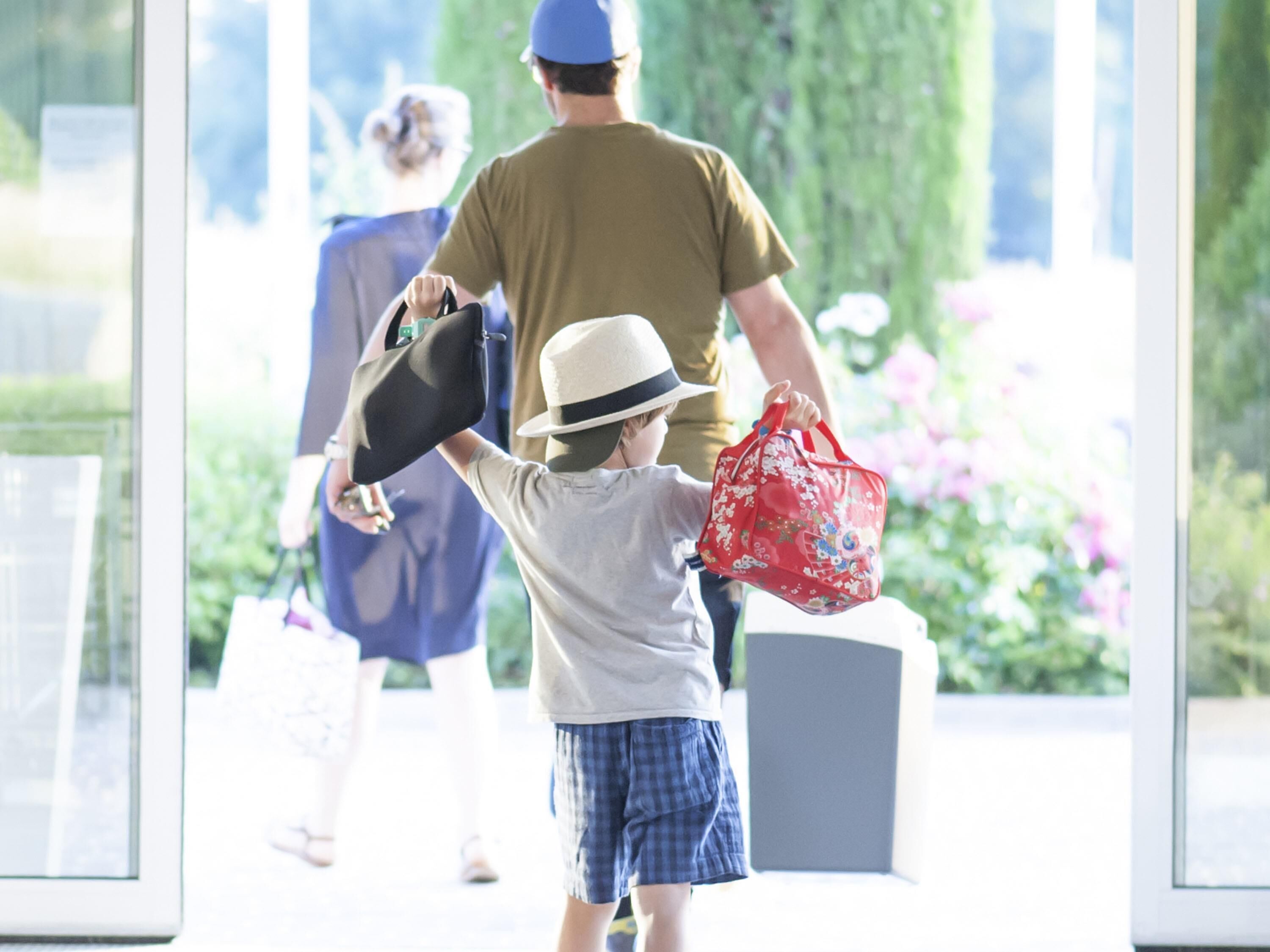 A child walking with a hat and a red bag, an adult holding a black bag, and another adult in the background.