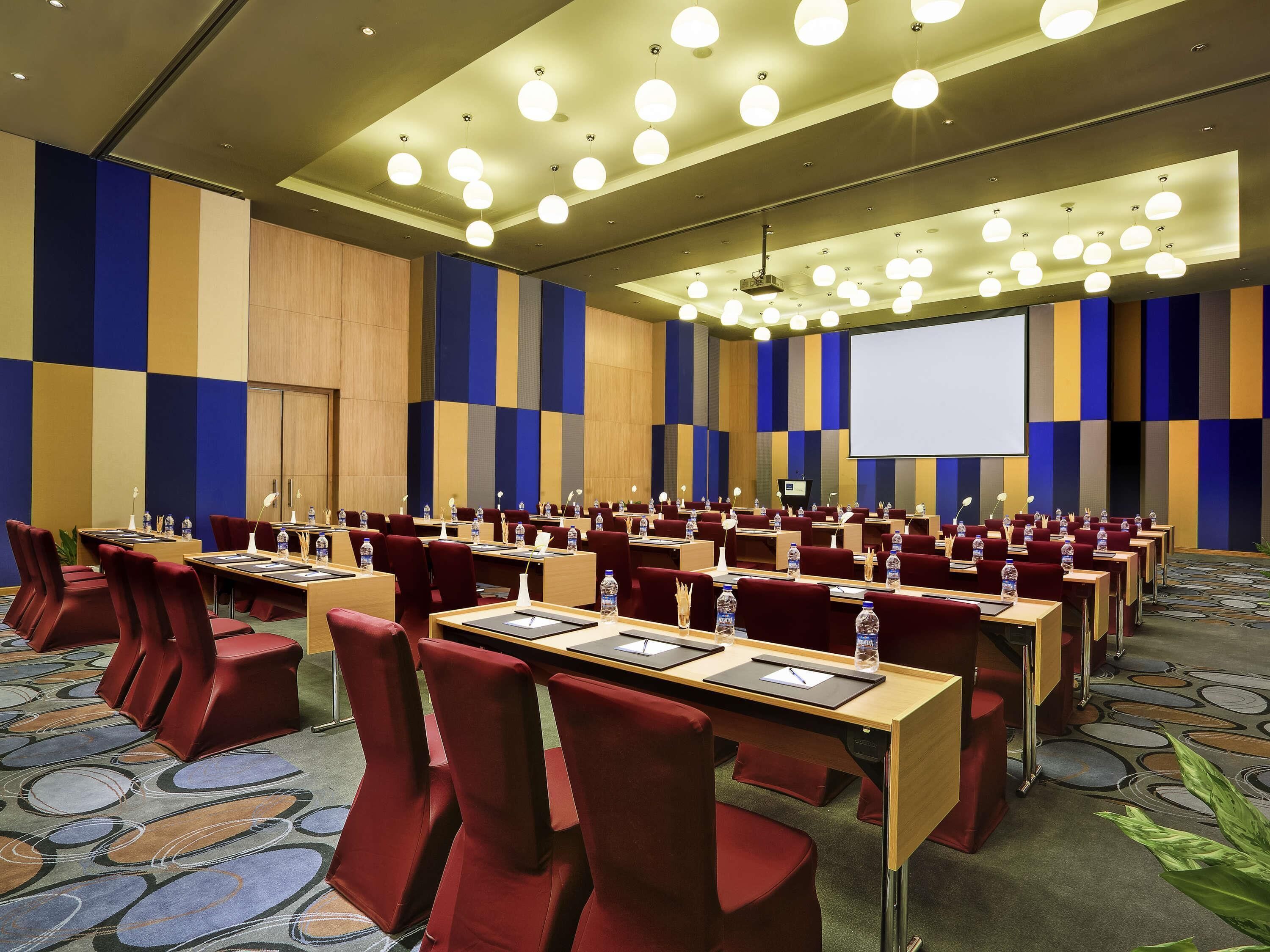 A conference room with rows of tables, chairs, and a large screen.