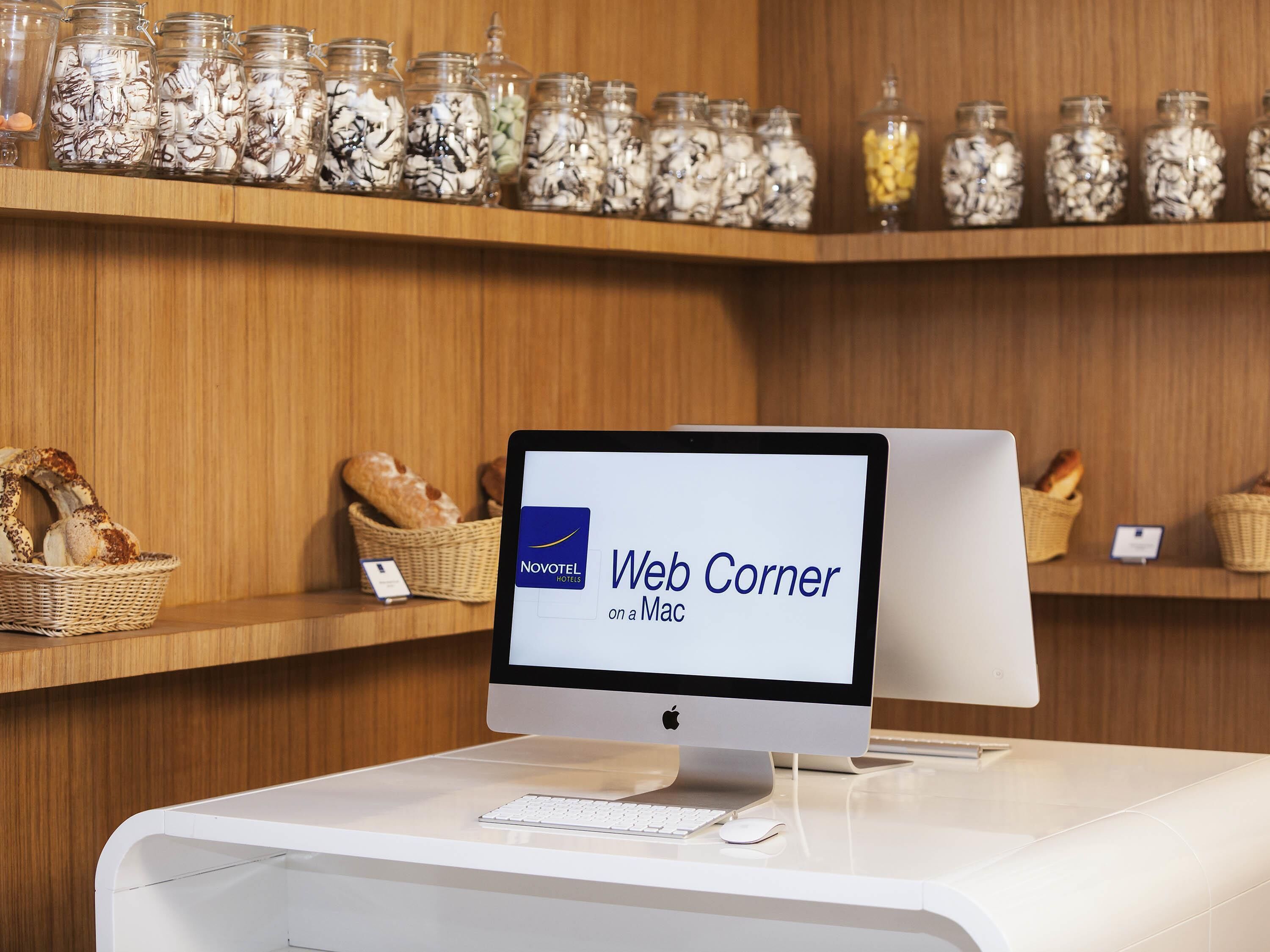 Uma área de recepção de hotel com um monitor de computador exibindo 'Web Corner on a Mac', cestos de vime com pão e prateleiras com potes de lanches.