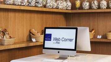 Uma área de recepção de hotel com um monitor de computador exibindo 'Web Corner on a Mac', cestos de vime com pão e prateleiras com potes de lanches.