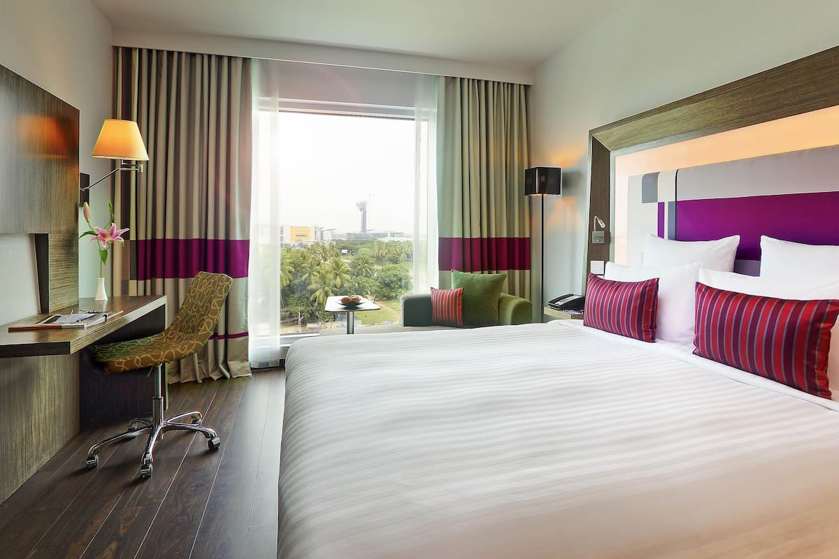 Habitación de hotel con una cama grande, un escritorio, una silla, una lámpara y vistas a la ciudad.