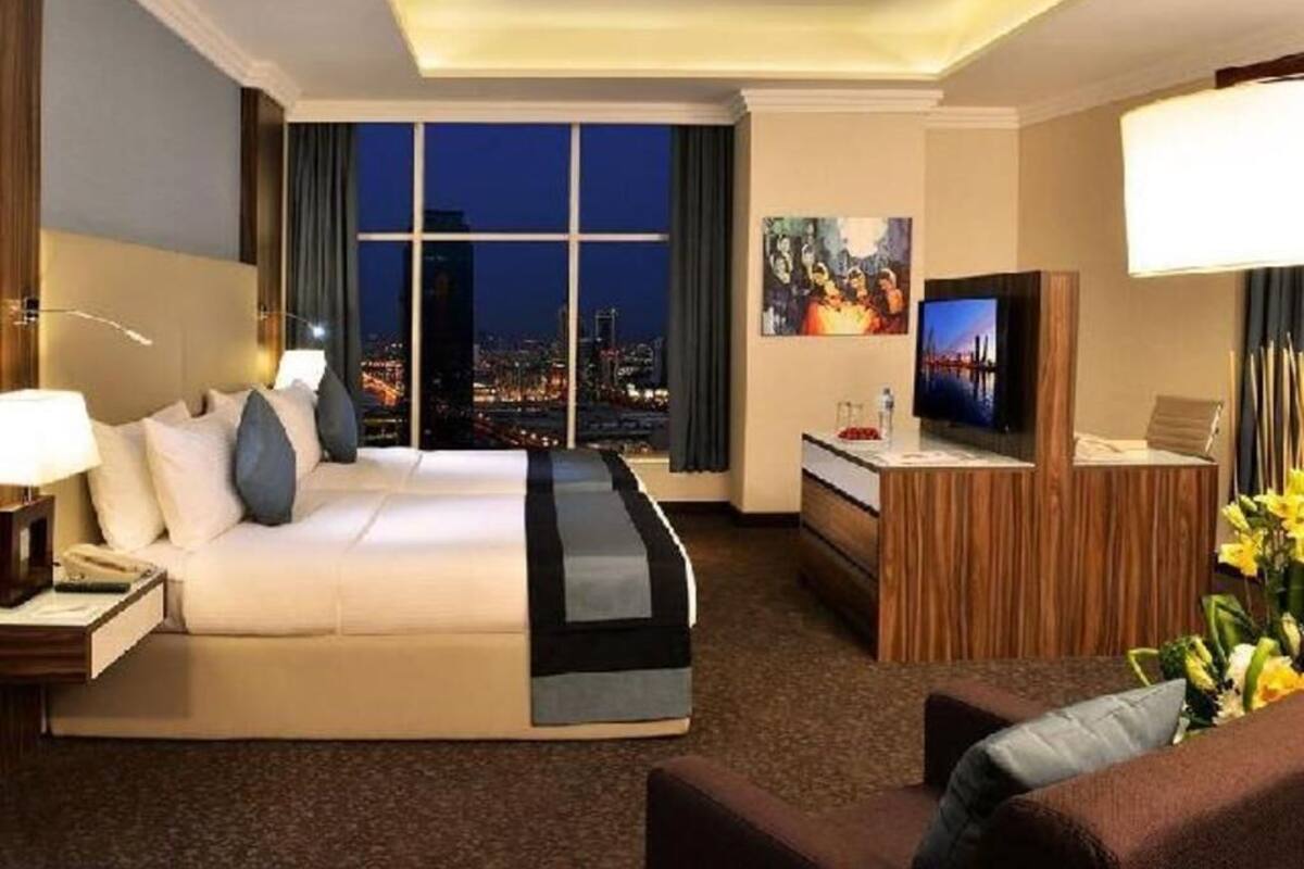 A hotel room with a bed, a desk, a TV, and a view of a cityscape at night.