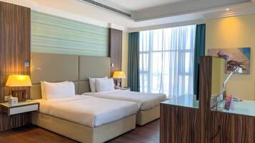 A hotel room with two beds, wooden flooring, a large window with curtains, and a desk with a lamp.