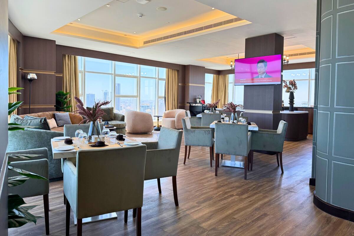 A spacious hotel lobby with a modern design, featuring a large seating area with sofas and chairs, a dining table, and a television screen displaying a news program.