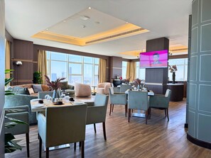 A spacious hotel lobby with a modern design, featuring a large seating area with sofas and chairs, a dining table, and a television screen displaying a news program. - Swiss-Belhotel Seef Bahrain (Manama)
