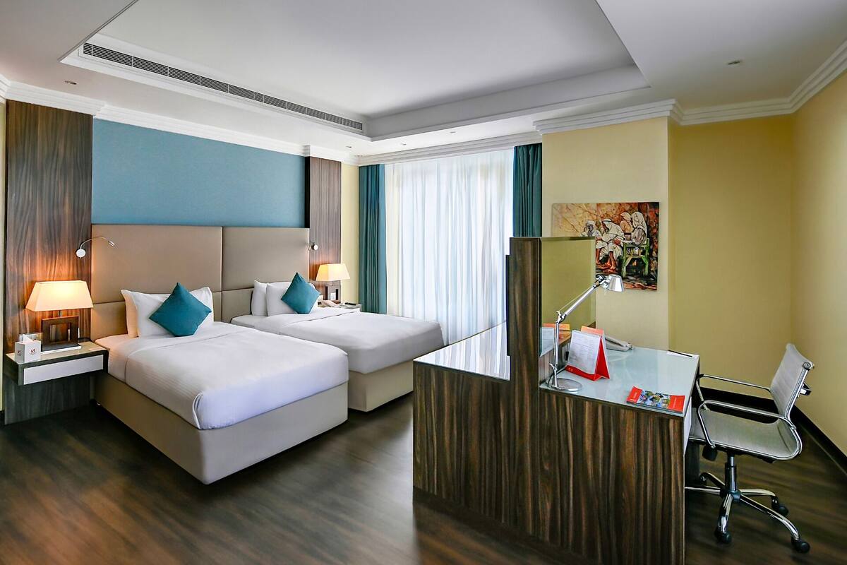 A hotel room with two beds, a desk, a chair, a nightstand, a lamp, and a painting on the wall.