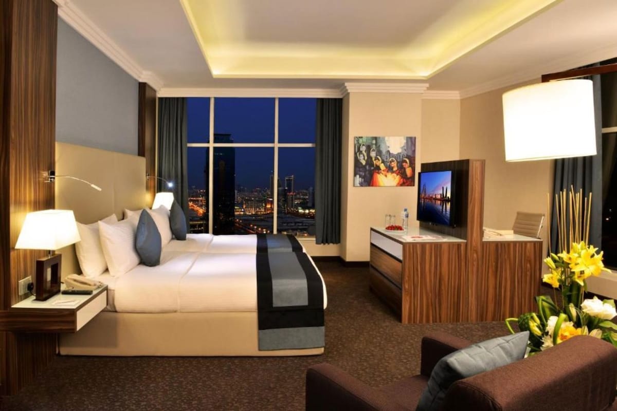 Ein Hotelzimmer mit einem großen Bett, einem Schreibtisch, einem Fernseher und Blick auf die beleuchtete Stadt.