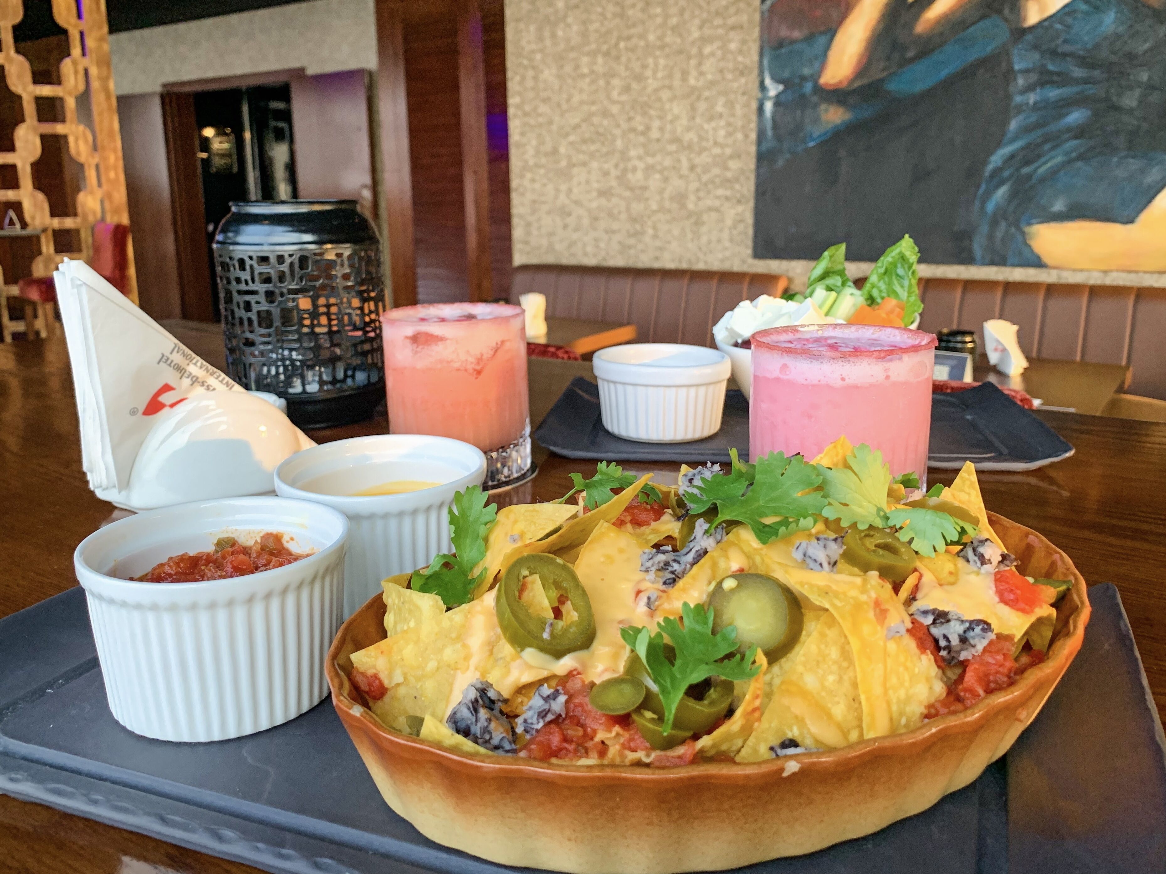 Un tazón de nachos con ingredientes, tres tazones pequeños de salsas, una vela y una bebida sobre una mesa.