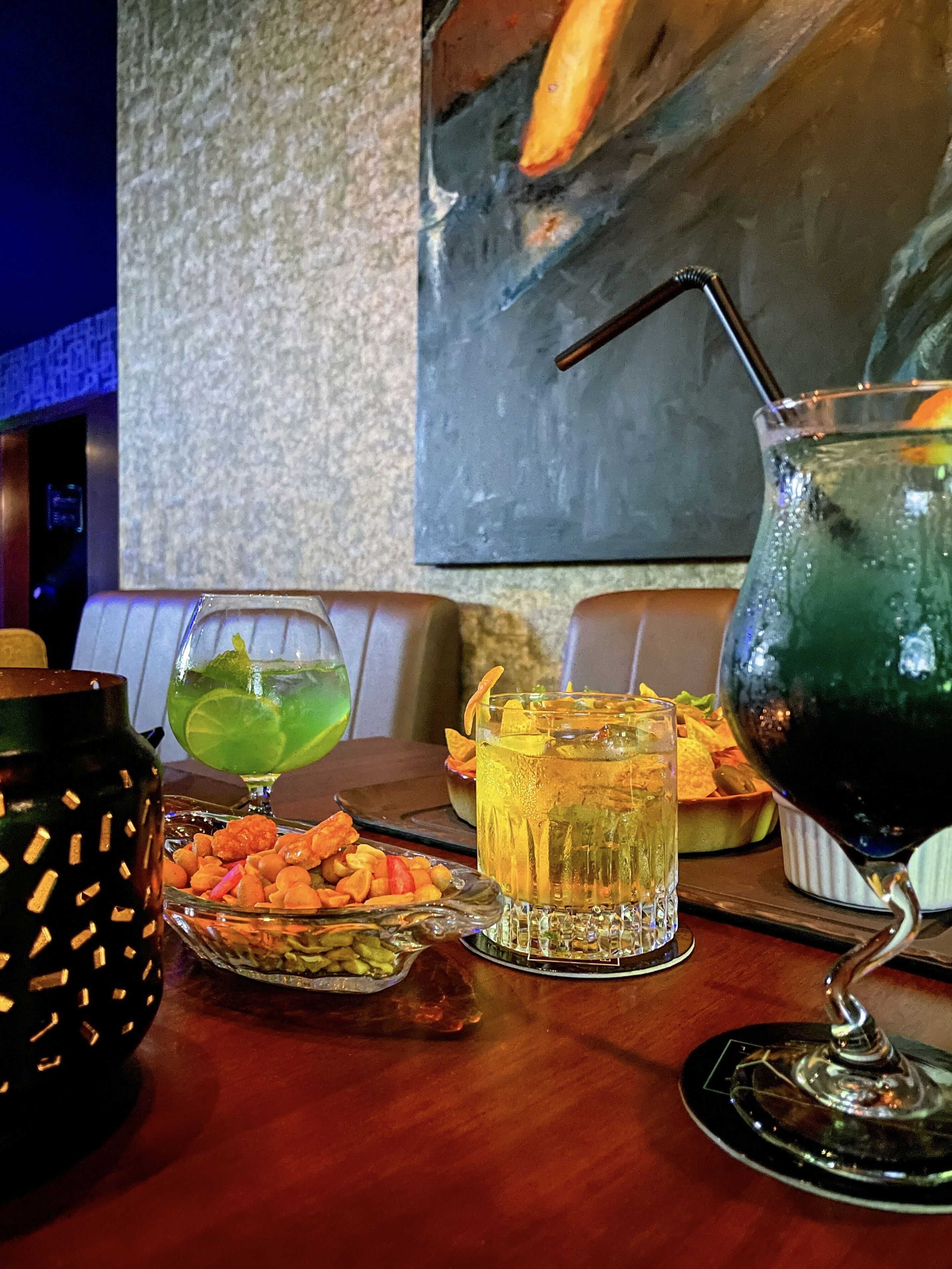 A bar area with a variety of drinks, including a green cocktail, a yellow drink, and a dark green drink with a straw. There are also snacks and a decorative candle on the table.